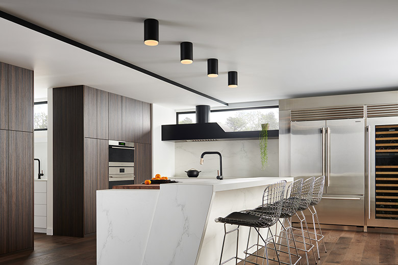 Interior modern kitchen with wide wood plank flooring, oversized marble kitchen island with a bowl of oranges on it and sink with black faucet, four chrome steel counter stools with black padded seating, walnut cabinets, high-end stainless-steel appliances, and a white ceiling where four Hinkley Lighting Cedric 30071BK extra small flush mounts are mounted over the island