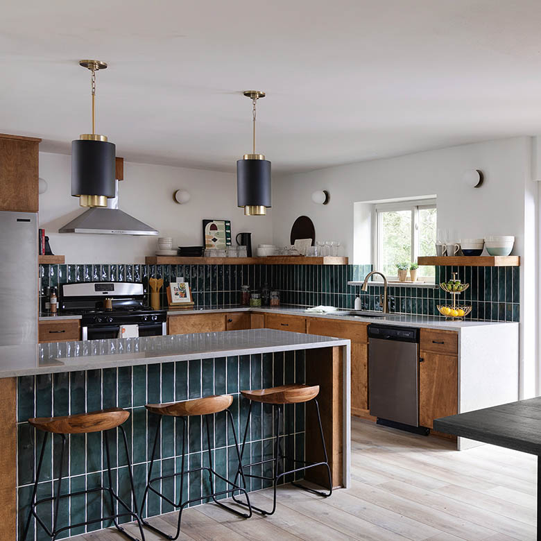Kitchen of mid-century modern home with bleached wood plank floor, white waterfall kitchen island with green tile faces, three black metal legged stools with wooden seats, natural oak cabinets with stainless-steel appliances, white waterfall countertop, sink has brass fixtures, backsplash in corner kitchen is the same green tile as the island, open wood shelving above the backsplash, white walls on which are mounted four Quorum Lighting 339-1-80 wall mount lights