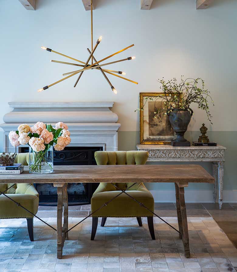 Eclectic dining room with slate tile floor and inlaid travertine tile center, with natural wood table and two pale green button-tufted upholstered chairs, marble finished sofa table on which sits a book, a large urn-shaped dark gray vase with flowers and a small painting with gold flecked frame, sage green half-wall with white above, large white fireplace, exposed beam ceiling from which hangs a Hinkley Lighting Archer 4766HB chandelier