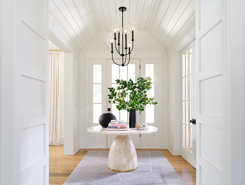 Residential entryway with wide plank hardwood flooring, area rug depicting concrete stones, marble mosaic table with brass-rimmed pedestal base and round mosaic top on which sits books, a vase with green branches, a small plate and an urn, white walls, white French doors and white wood ceiling from which is hung a Hinkley Lighting alchemy 20525BK chandelier