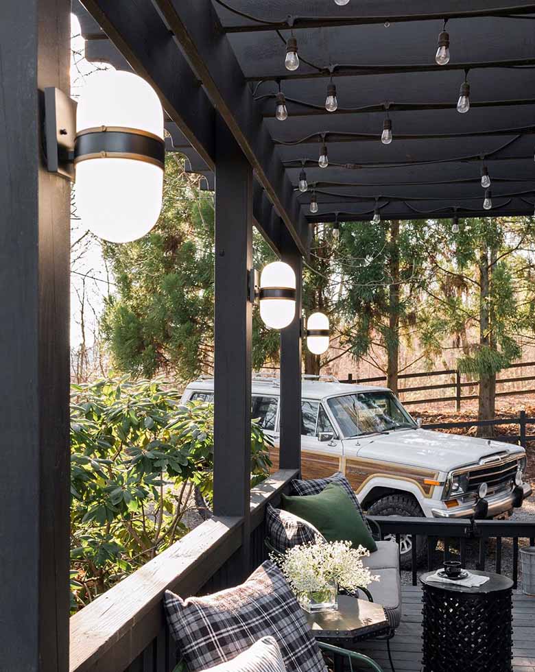 Outdoor patio scene with wood deck and trees and older jeep Grand Cherokee in the background, wood railings, green metal furniture with gray striped cushions, various colored throw pillows, black wood pillars on which are hung three Crystorama Lighting Capsule CAP-8501 outdoor wall sconces, black painted wood pergola from which are hung multiple sets of string lights