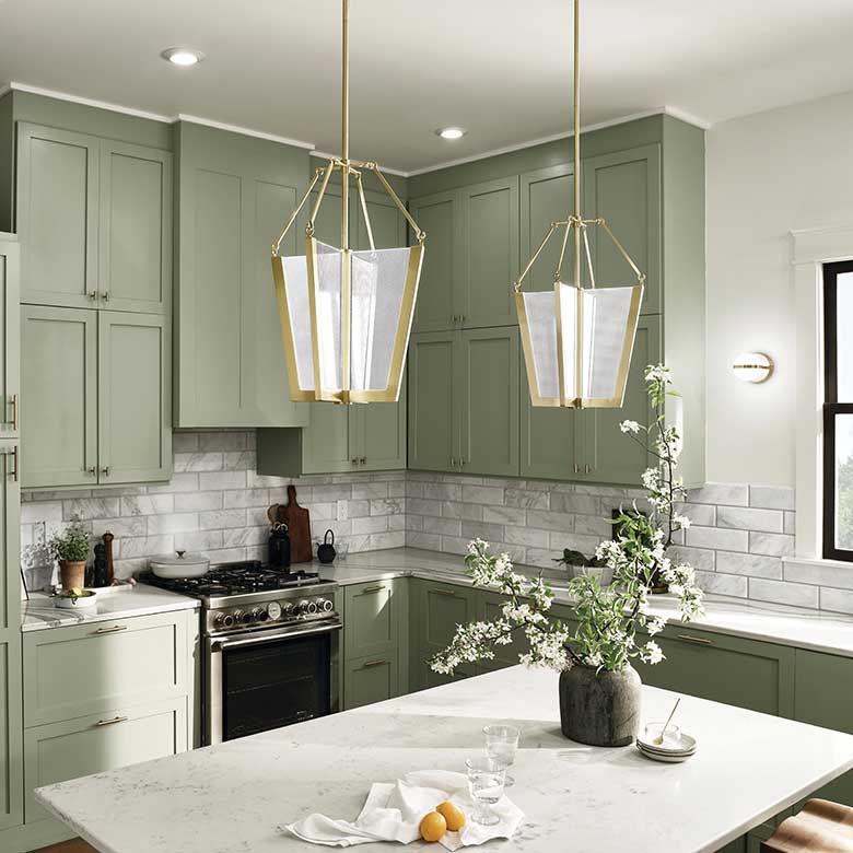 Interior kitchen show with hardwood floors, green cabinets and kitchen island both with white marbled tops, stainless-steel appliances, marble backsplash, white walls leading to a white ceiling from which hangs two Kichler Lighting Calters 5229CGLED pendant lights