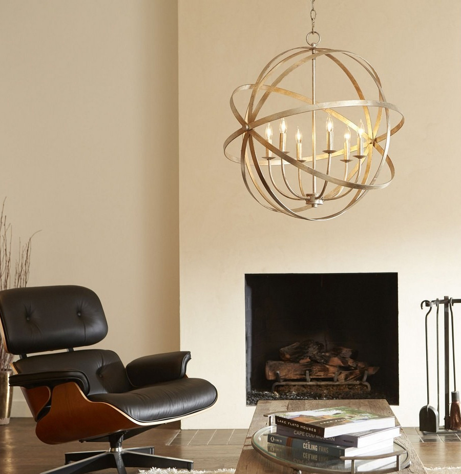 Modern living room application shot with wood table, fireplace, cream-colored walls, black Eames lounge chair and six-light orbital chandelier in an aged brass finish