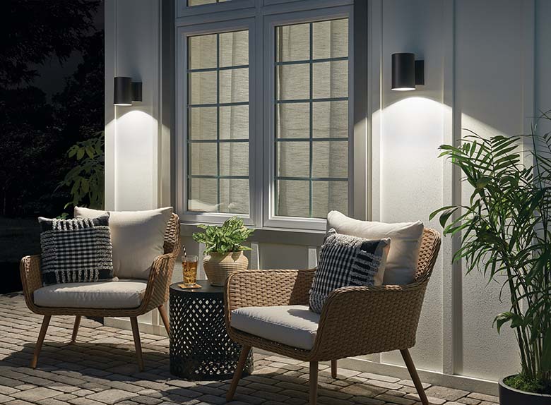 Outdoor patio scene with cobblestone pavers, two natural rattan chairs with off-white cushions, a black metal basketweave side table on which sits a potted plant and glass of iced tea, white board and batten walls and two Kichler Lighting 11309BKTLED outdoor wall sconces flanking a white window with green trim