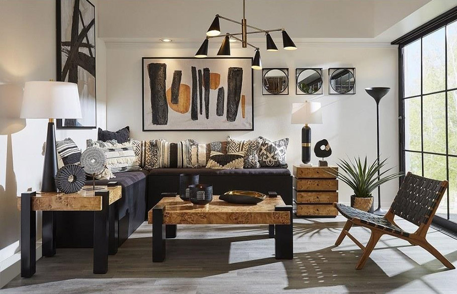 Interior modern living room with gray plank flooring, black upholstered furniture, black and natural wood coffee and end tables, white walls on which are multiple black and white paintings and three small mirrors, and white ceiling from which hangs an Elk Home Calder 89217/6 modern chandelier