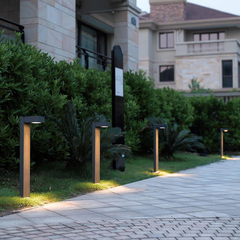 Outdoor walkway with brick and square stone pavers, in high end commercial setting with buildings of stone tile, lush green shrubbery, and four Eurofase Lighting 31909-027 LED bollard path lights lining the walkway