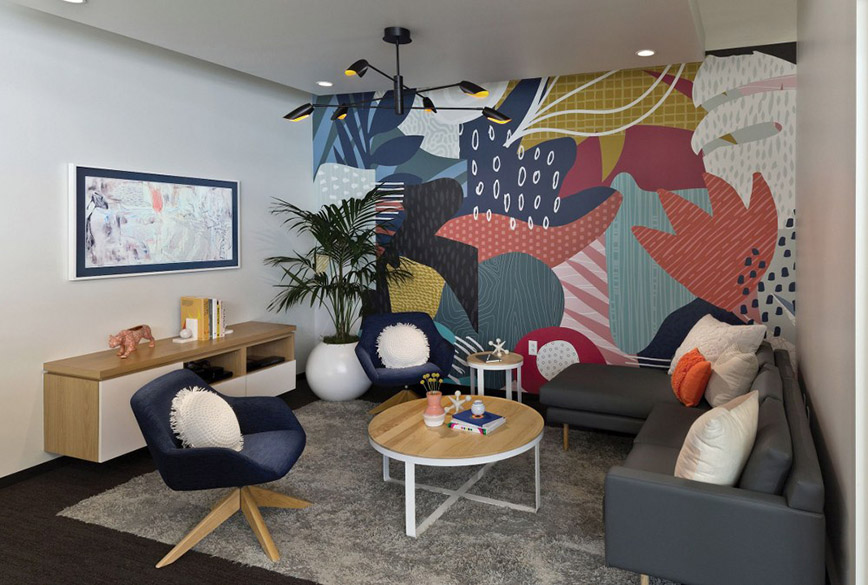 Modern living room with dark wood floors, gray shag area rug, gray leather modern sofa with built-in chaise lounge, white metal end and coffee table with natural wood tops, two blue velvet side chairs, wood credenza, large plant potted in white ball planter, white wall with modern horizontal painting, accent wall with multiple colors and patterns, white ceiling from which hangs a Fredrick Ramond Bowery FR35806BLK multi-tier modern chandelier