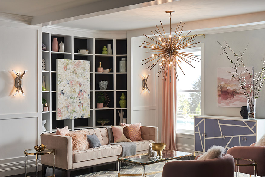 Interior of midcentury modern living room, with dark wood floors, gray zig-zag pattern area rug, brown tufted upholstered sofa, mauve suede side chairs, gold leaf coffee table and end table, white dresser with painted dusk blue geometric patterns on the front on which sits a vase with cats tails in it, white board and batten walls with curved built-in bookcase, white ceiling with painted white exposed beams with a Fredrick Ramond Lighting Tryst FR43015BNG stem hung pendant in burnished gold finish