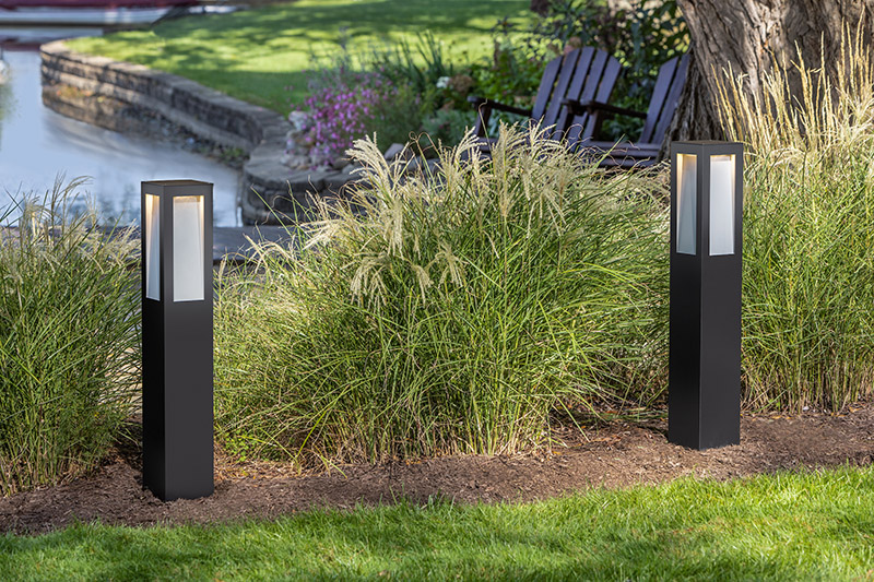 Outdoor scene around man-made pond, with stone architectural pond lining, tall grass shrubbery, wooden Adirondack chairs, and pathway with two Hinkley Lighting Taper 15288BZ LED bollard path lights