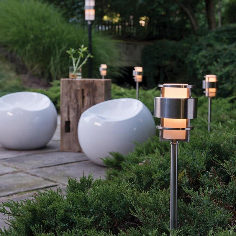 Outdoor walkway with lush green shrubbery, white plastic modern chairs with a natural wood table, and four Hinkley Lighting Saturn 1579SS-LL modern path lights