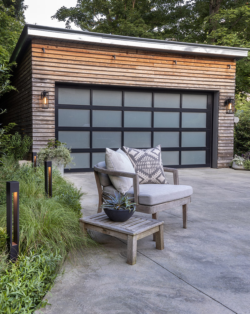 outdoor scene of detached garage with textured concrete driveway flanked by Hinkley Atlantis 15602 large bollard lights, washed wood furniture, deep foliage shrubbery, natural wood garage with two-car modern multi-panel frosted glass door with black frame