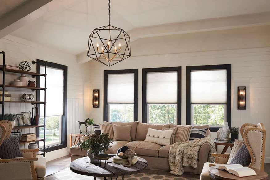 Interior living room with wide plank floors, large brown spotted area rug, brown upholstered sofa and tufted side chairs, wood spindle top coffee table, wooden end tables, white board and batten walls with black trim around multiple windows, white ceiling with white painted wood beams from which hangs a Hinkley Lighting Astrid 3025MM large chandelier