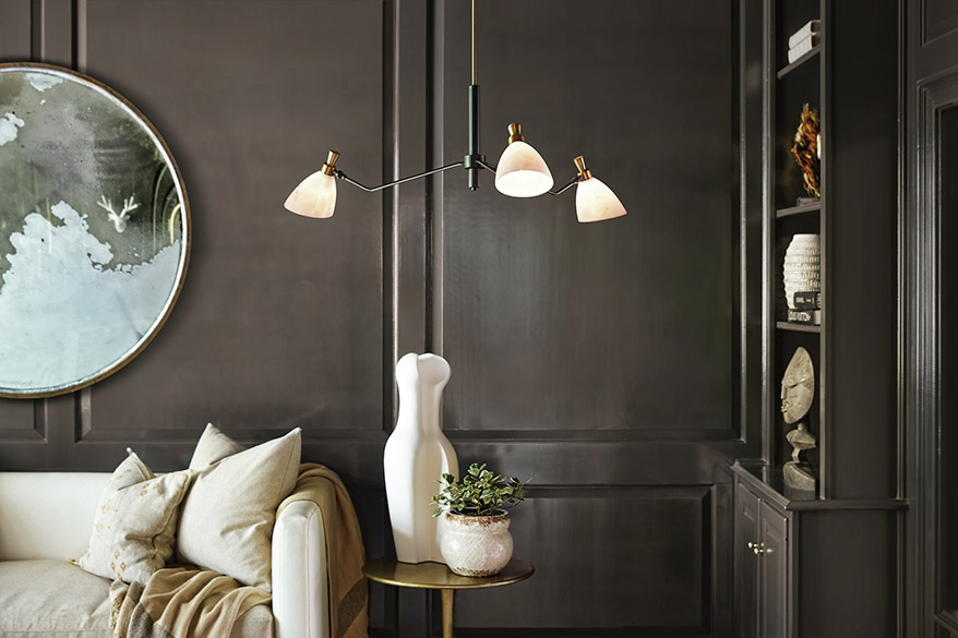 Interior living room with dark mocha stained wood walls, light taupe sofa and cushions, gold end table, built-in mocha wood bookcase, large circular mirror, and from the ceiling hangs a Hinkley Lighting Sinclair 33793HB LED chandelier