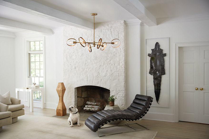 Interior of a modern living room, with light wood plank flooring, large beige throw rug, beige upholstered furniture, natural alligator hides chaise lounge, white walls upon which is an alligator hide, and stone fireplace painted white, with white ceiling with white-painted wood beans, from which hangs a Hinkley Lighting Desi 37296LCB large chandelier