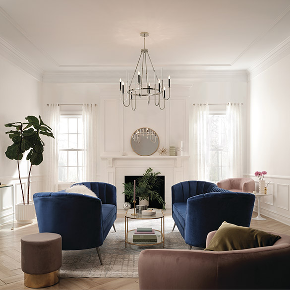 Modern living room with herringbone wood plank floors, gray patterned accent rug, blue upholstered couched set opposite each other with a gold coffee table in between, mauve side chairs and ottoman, potted plant in white pot, white fireplace surround, board and batten half-walls painted white, and white tray ceiling from which hangs a Kichler Lighting Kadas 52210PN large chandelier