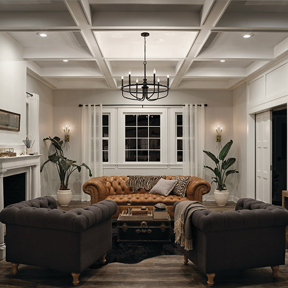Interior living room scene with wide plank floors, brown leather sofa, dark brown suede side chairs, steamer trunk as a coffee table, two potted plants, white marble fireplace surround, beige walls with large white windows and white coffered ceiling from which hangs a Kichler Lighting Capitol Hill 52304BK large chandelier