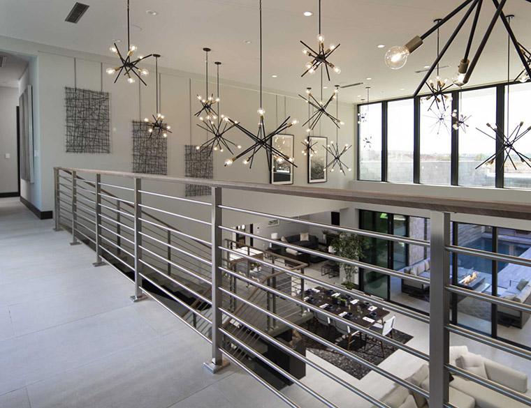 Open view of loft into downstairs are in modern household, showing tiled floors, beige furniture, white walls, gleaming steel railing on second floor, and multiple Progress Lighting Astra P400108-020 chandeliers hanging from various heights