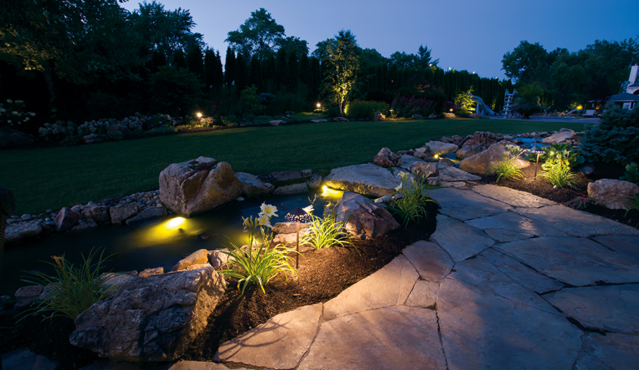 Outdoor landscaping scene with large stone pathway, dual coy ponds, lush green grass and the path lit by Kichler Lighting 15871 hammered roof path light