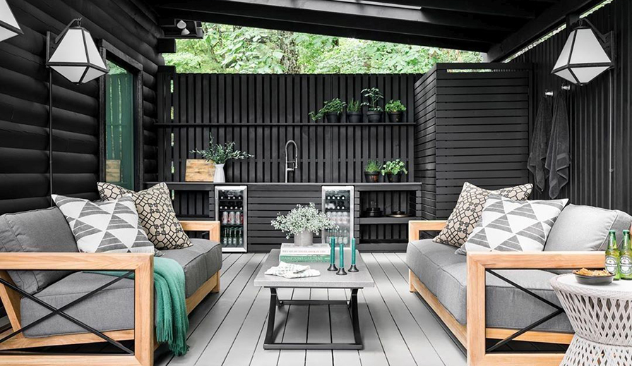 Outdoor covered patio scene with gray wood plank floor, small outdoor drink area with stainless steel refrigerators, charcoal wood walls from which hang multiple Crystorama Lighting Glacier GLA-9702-OP-BC outdoor wall lights, charcoal colored ceiling with skylights