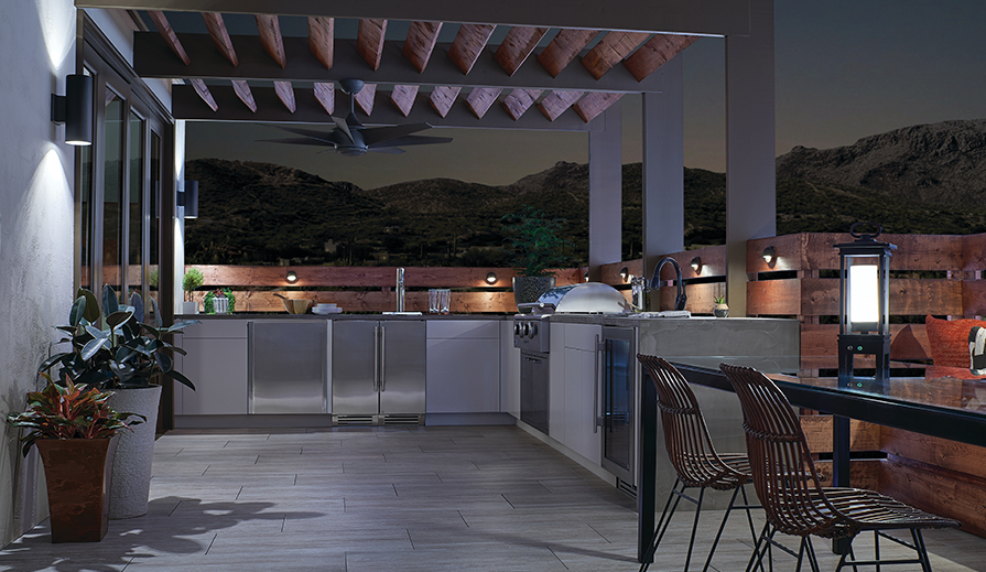 Outdoor kitchen at twilight overlooking mountain range with tiled floor, wood slatted railing with step-lights for counter illumination, white cabinets, stainless-steel appliances, gray countertop, black metal dining table with glass top on which sits a portable LED light, white textured walls on which hangs two Kichler Lighting 11310BKTLED outdoor wall lanterns, black floor-to-ceiling bifold doors, and black pergola over kitchen area with natural wood slats