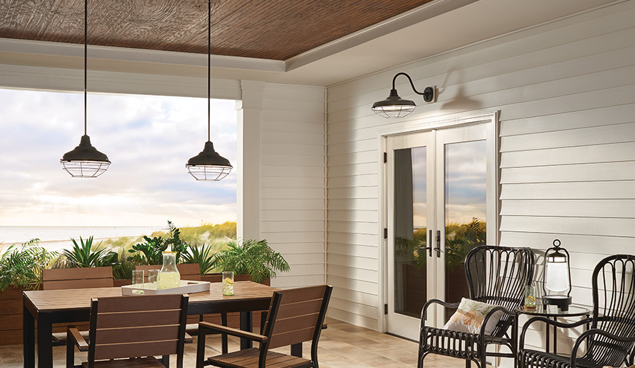 Patio scene overlooking the ocean with wood tile floor, black metal dining table and chairs with wood slats as seats, backs and tabletop, black metal side chairs flanking a black metal side table with a glass top, cream-colored walls, and full panel glass French doors over which is hung a Kichler Lighting Pier 49991BK gooseneck wall lamp, dark wood ceiling from which hangs two Kichler Lighting Pier 49992BK convertible pendants over the table
