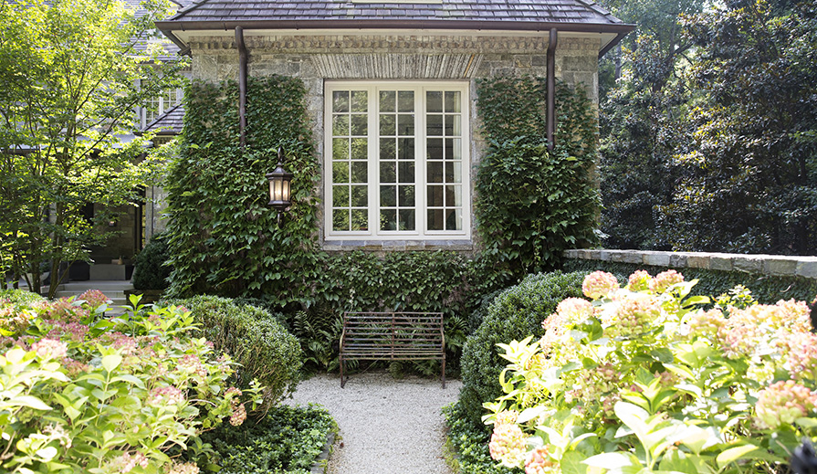 Outdoor garden scene looking at traditional home made of inlaid stone with garden paths of pebbles, lush greenery and flowers and at the intersection with left-turn ahead is a two-person brown metal garden bench, stacked stone half-wall next to it culminating in an exterior wall of the house covered in ivy from which hangs a single Hinkley Lighting Raley 1609 outdoor traditional wall lantern