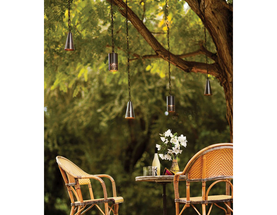 Outdoor yard scene with blurred trees in the background, with sand scaped dirt floors, rattan bar-height stools, bar-height small round table with flowers and drinks upon it, and from a tree branch above the table hangs multiple Hinkley lighting Calder 1511BZ conical lights and Hinkley Lighting Calder 1512BZ cylindrical lights