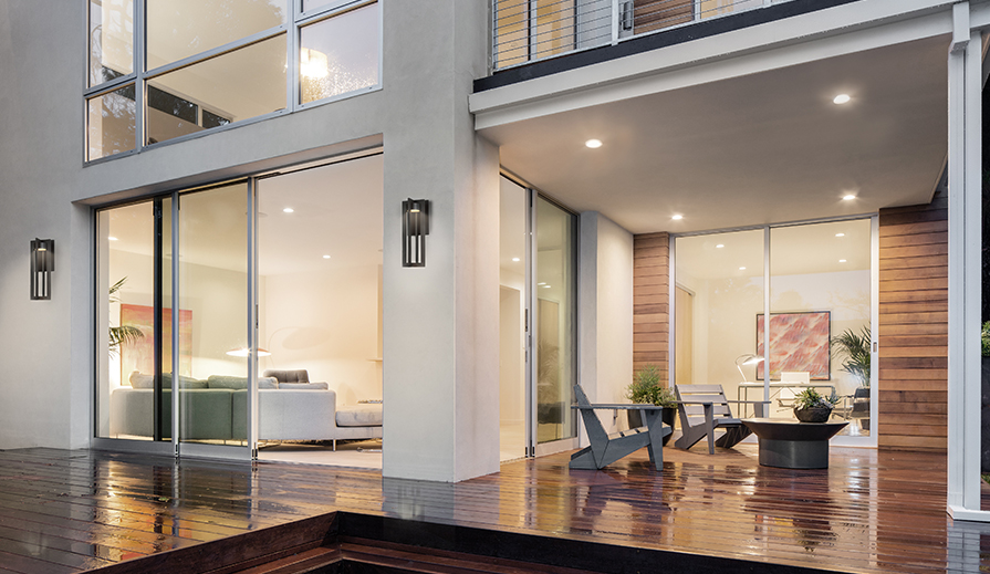 exterior view of patio on contemporary home, with finished wood plank decking, gray wood modern Adirondack chairs and matching coffee table, with floor-to-ceiling panel windows on all floors, and white textured walls on which are hung multiple DweLED chamber WS-W48625BK outdoor wall-mount lights