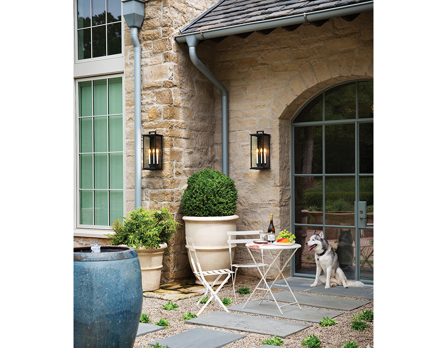 Outdoor patio with pebble flooring and inlaid stepping stones, potted plants, stone house on which are hung multiple Hinkley Lighting Weymouth 20015BK outdoor wall lanterns