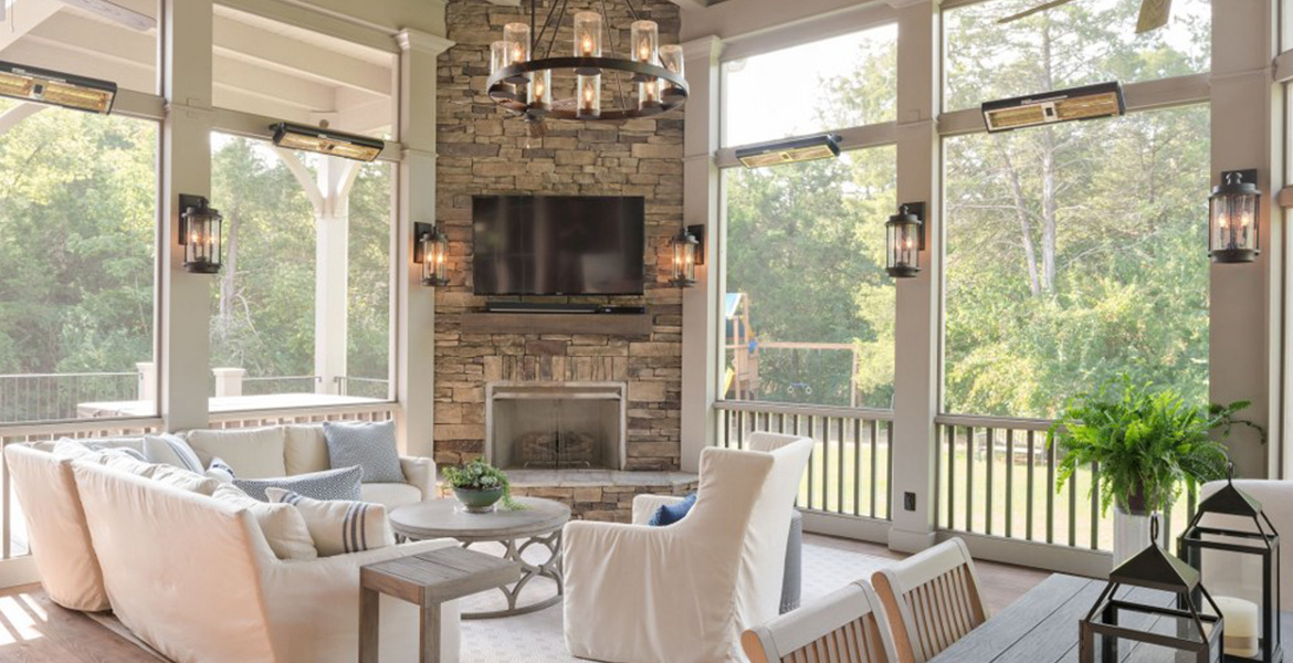 Upscale screened patio shot with pine floors, white geometric patterned area rug on which sits white cloth covered patio furniture, outdoor dining table, stacked stone fireplace with television mounted above it, white walls with floor-to-ceiling windows, large outdoor chandelier and four Solaira Alpha SALPHA-4000 outdoor winter heaters in black